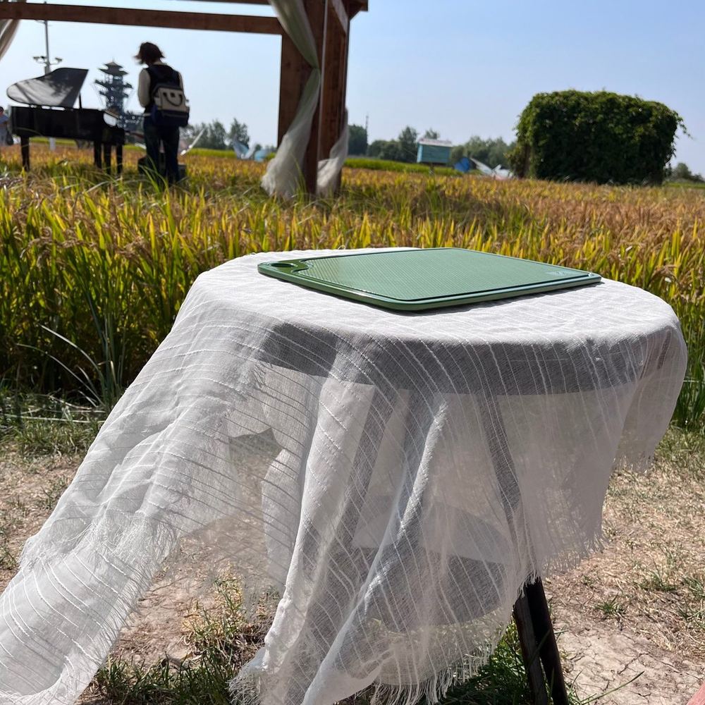 Green rice husk chopping board on a linen-covered table set up in a rice field — product photography at harvest season