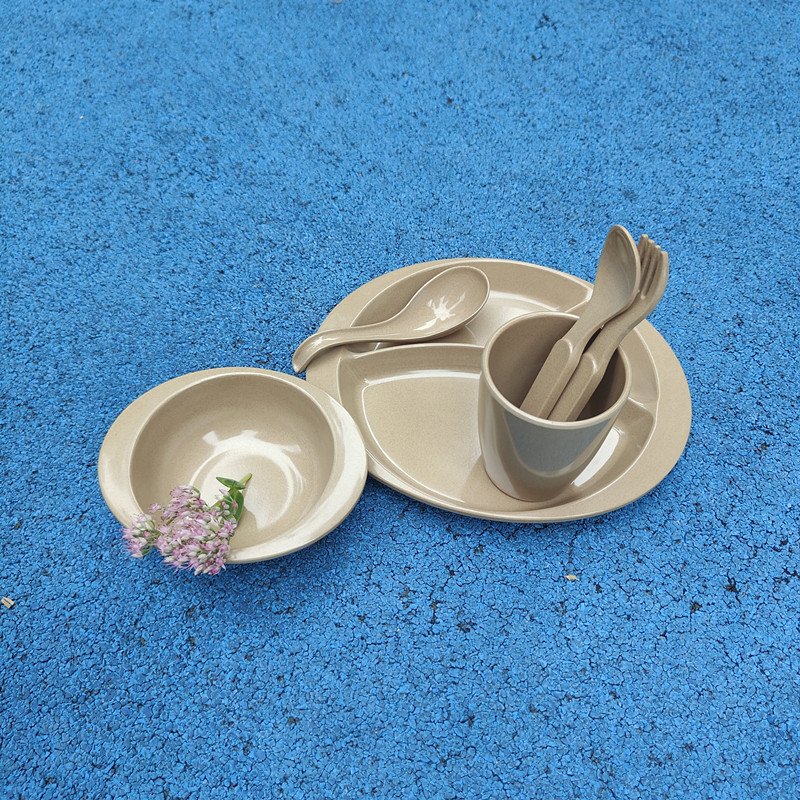 Six-piece rice husk kids tableware set — divided plate, snack bowl, drinking cup, fork, spoon and soup spoon in natural beige — on a playground surface